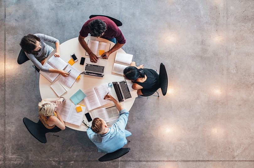 Five people sitting around a table. Each person is using a digital device. They are working together on a project.